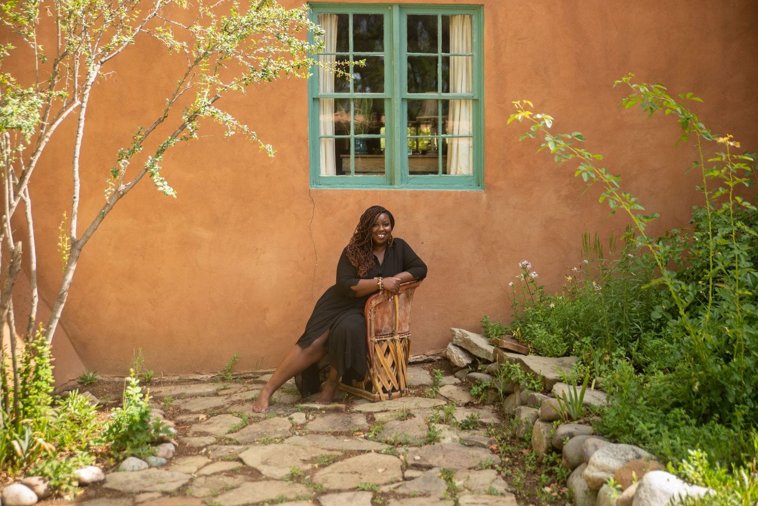 Kali Shelton sitting on a wooden bench in a black dress with braids, in a garden setting