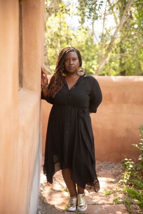 Kali Shelton leaning against a sunlit adobe wall in a black dress with braids
